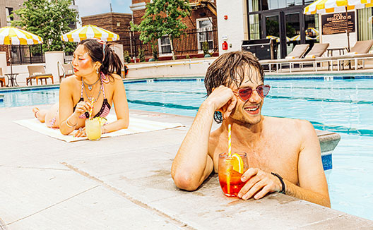 Man in Cottonwood pool with cocktail, woman laying on pool desk on tool with cocktail